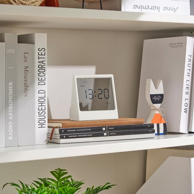 A modern white RÖDBENA digital clock sits on a stack of books on a shelf, surrounded by decor items. It displays time and temperature.