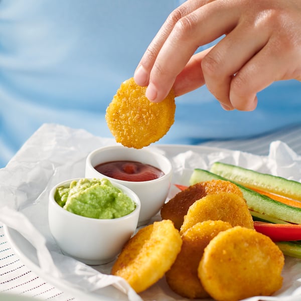 Hand holding golden-brown breaded wheat piece over a tray with more pieces, vegetable sticks, and two dipping sauces.