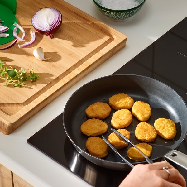 Person cooking breaded wheat-based pieces in a black frying pan on a stove. A wooden cutting board nearby has sliced onions, garlic, and herbs.