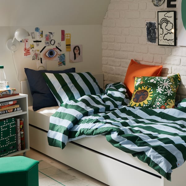 Cosy bedroom with green-white striped bed, colourful pillows, books, and lamp.