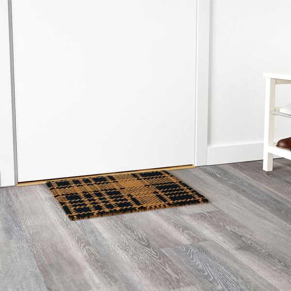 Rectangular indoor mat, black/beige chequered, placed against white door/wall, grey floor.