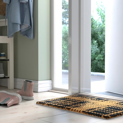 Yellow doormat, named SLIPRAR, with black details and bold check pattern welcomes guests by a large window with pink trainers.