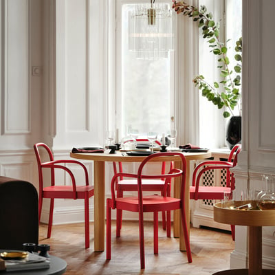 Modern dining area with red, curved STOCKHOLM 2025 chairs around a light wood table, under a chandelier, beside a large window. Bright and spacious.