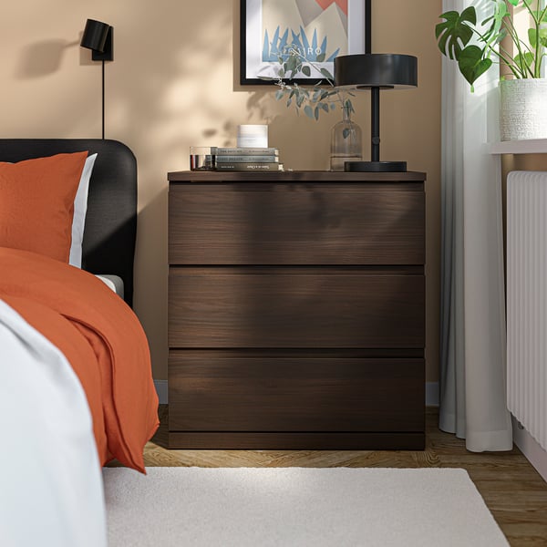 Dark wooden STORKLINTA bedside chest; includes drawers, lamp, books, and vase. Positioned beside bed, smaller than bed.