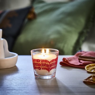 A small, lit red & white VINTERFINT candle sits on a table, surrounded by a napkin, bowl, and citrus slices. The glass cup displays a floral pattern.