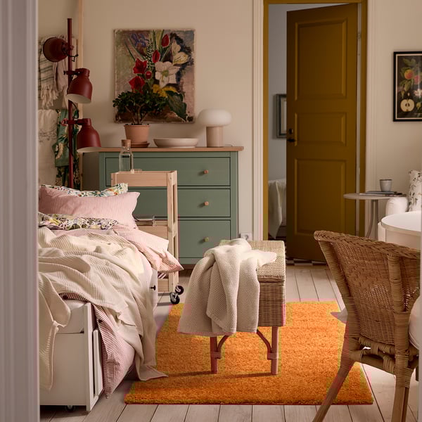 Bright bedroom with an orange arende rug, wicker chair, white bedding, green dresser, and floral art. Soft, thick rug contrasts cosy decor.