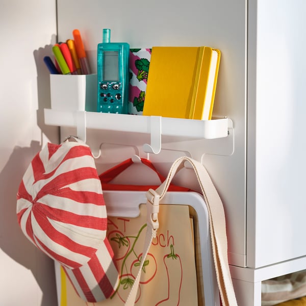 White hook rack with shelf and container hangs on cabinet, holding colourful items like a striped bag, notebook, and markers.