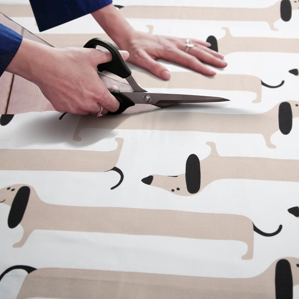 Person holds scissors cutting playful dog-patterned fabric, likely for sewing or crafting.