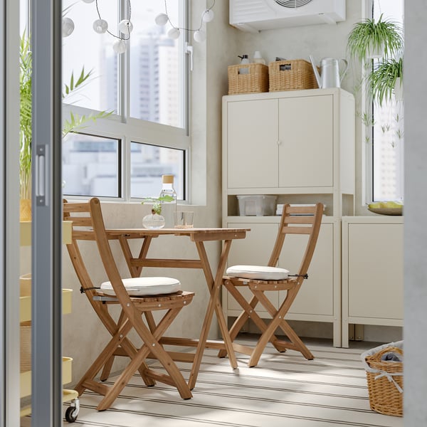 Compact ASKHOLMEN citrus wood dining set with cushions near a white cabinet and window.