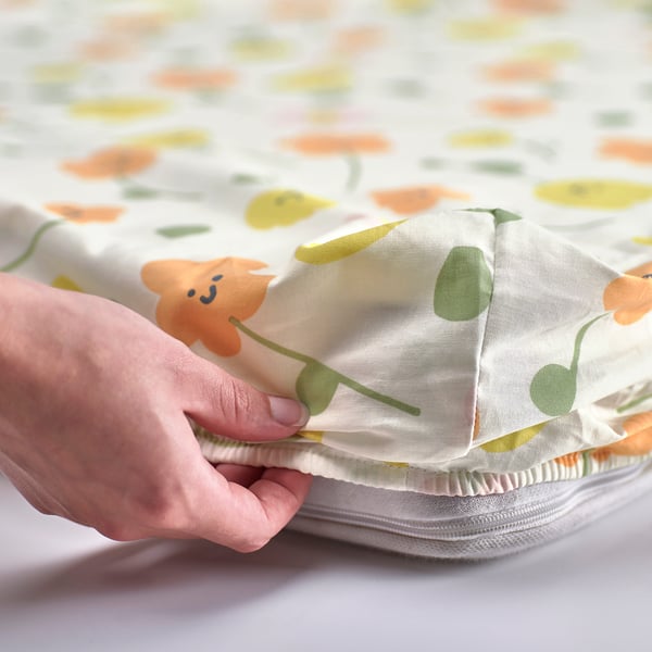 A hand holds a corner of a fitted sheet with cheerful flower prints, showing elastic edges, designed to fit mattresses up to 10 cm thick.