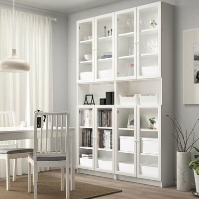 White BILLY bookcase with glass doors, holding books and decor beside dining area.