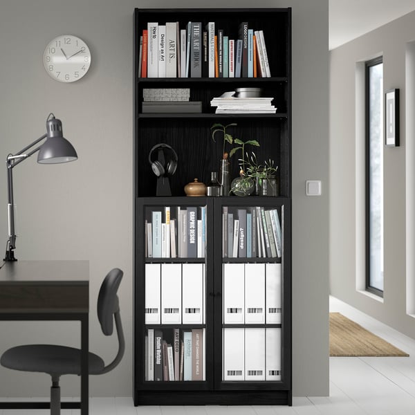 Black BILLY bookcase with books & decor, tall above desk & chair, glass doors, wall clock, lamp.