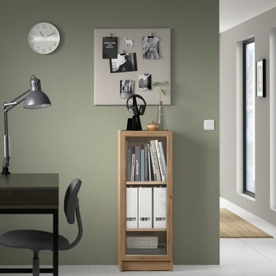 Home office with BILLY bookcase in wood tone, black chair, desk lamp, and bulletin board on green wall. Clock above.