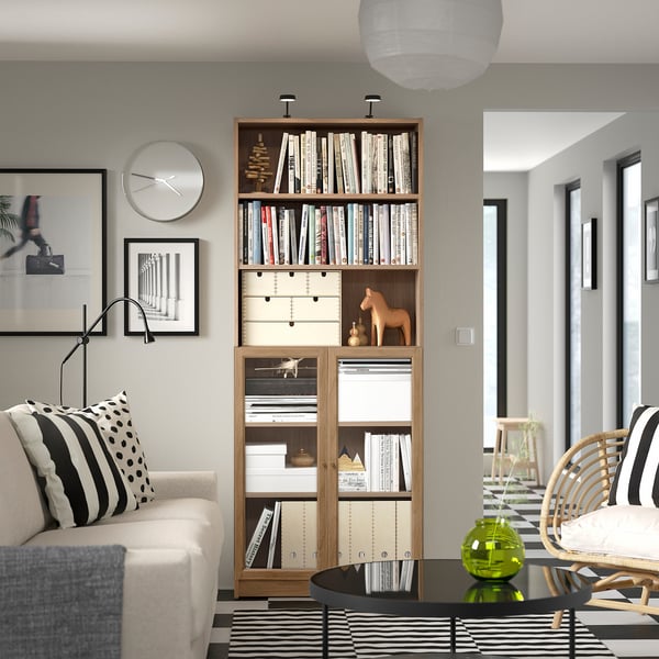 Modern living room with BILLY bookcase, beige couch, tall clock, art, rattan chair, and green vase.