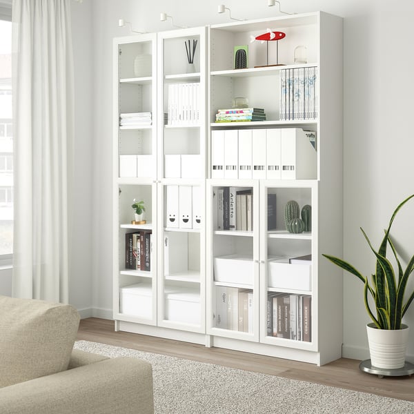 White BILLY bookcase with glass doors, books, binders, plants. Beside, beige armchair and potted plant. Bright room with light walls.