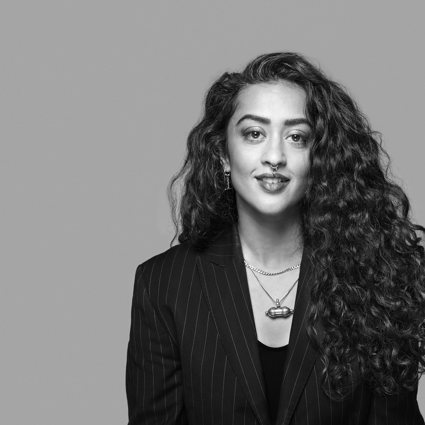 Black and white portrait of Akanksha Deo wearing a striped blazer with long curly hair, smiling softly.