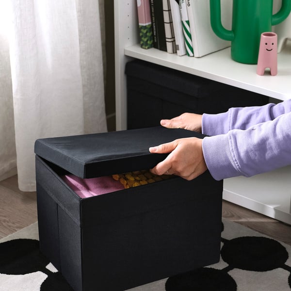 Person lifting lid of black DRÖNA storage box, revealing contents inside. Box sits on patterned rug, with white bookshelf in background.