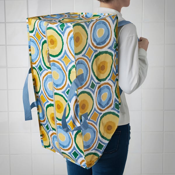 Person carrying colourful, foldable storage bag with handles and shoulder straps against tiled wall.