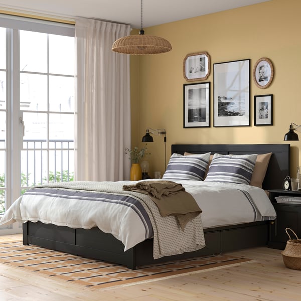 Modern bedroom with black oak bed, striped linens, storage boxes, large window, and artwork.
