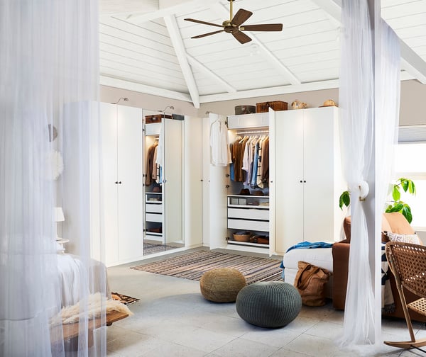 Bright, white closet with mirrored door, next to bed in airy room, light wooden floor, two round pouffes at foot.