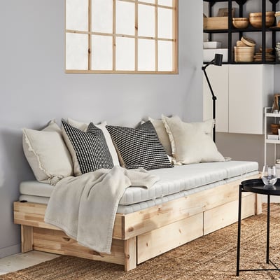 GLAMBERGET extendable day-bed in light wood with pillows, under a window, beside a table, in front of cabinets.