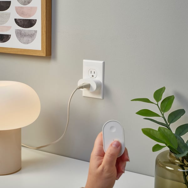 Hand holds remote near smart plug, controlling lamp, green plant visible.