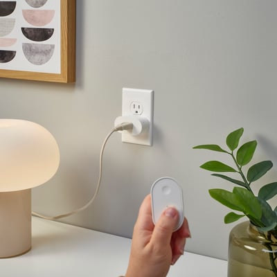 Hand holds remote near smart plug, controlling lamp, green plant visible.