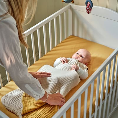 Blonde person adjusting sleep bag on baby in white crib, ensuring comfort with soft organic cotton.