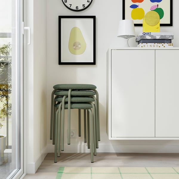 A modern, minimalist room with a stack of green stools, a clock, a plant, colourful art, and a white cabinet with games on top.