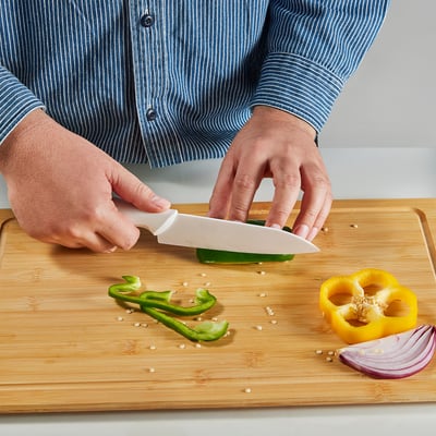 A cutting board with vegetables and a ceramic knife. The knife, labelled HACKIG, has a sharp edge for slicing, and its handle has an anti-slip grip.
