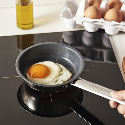 A hand holds a small frying pan on a stove, cooking a fried egg. The pans non-stick surface allows for easy cooking.