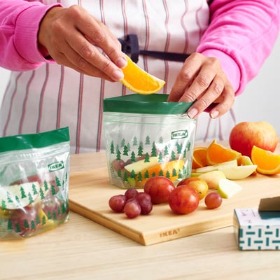 A person sealing an orange slice in a green IKEA ISTAD bag on a wooden board, showing assorted fruits inside.