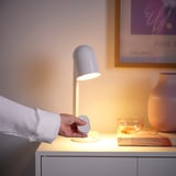 A hand turns on a white lamp on a bedside table, with a remote, vase, and photo frame nearby.