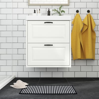 Modern bathroom with white cabinet and tiles, mustard towels on black hooks, black & white bath mat.