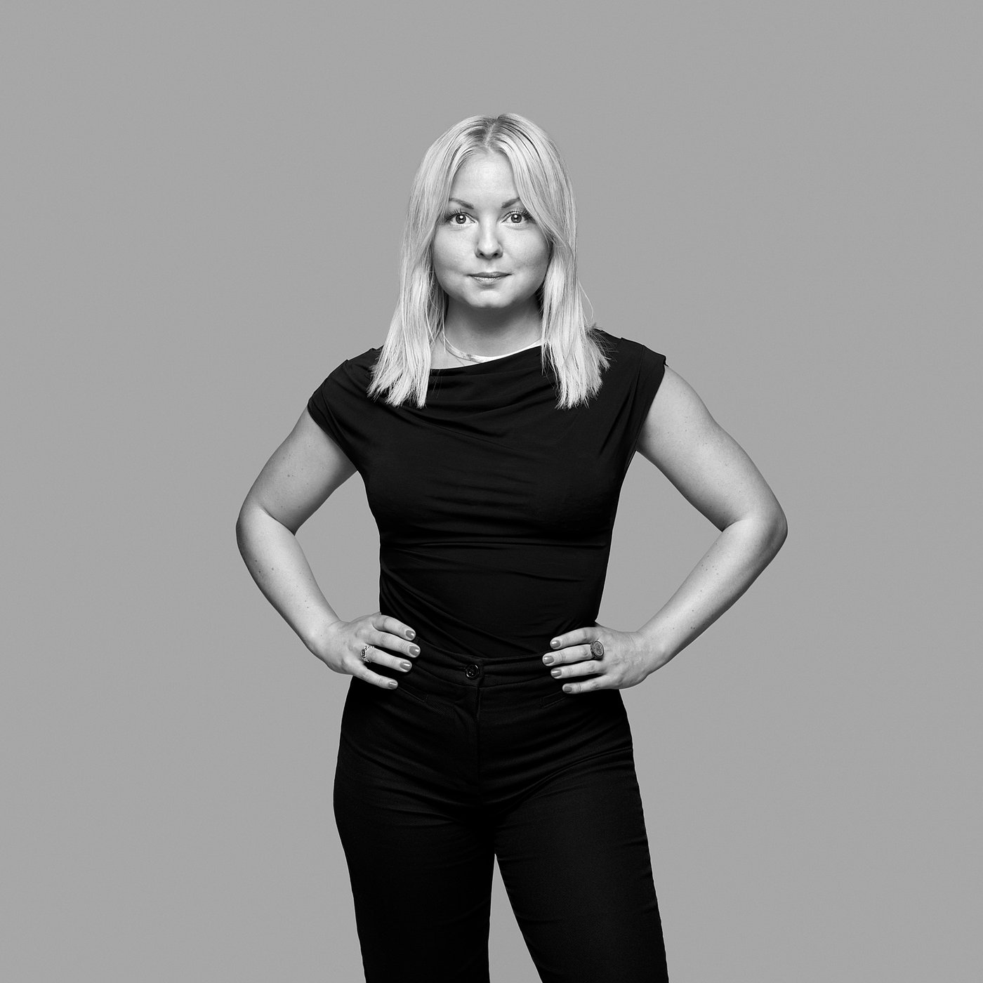 Black and white portrait of the designer posing confidently with hands on hips, wearing dark clothing.