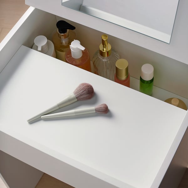 White dressing table with open drawer, makeup brushes inside, surrounded by various cosmetic bottles.