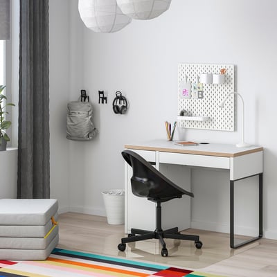 Modern home office with white MICKE desk, black chair, and pegboard. Bright rug, grey curtains, and plants.