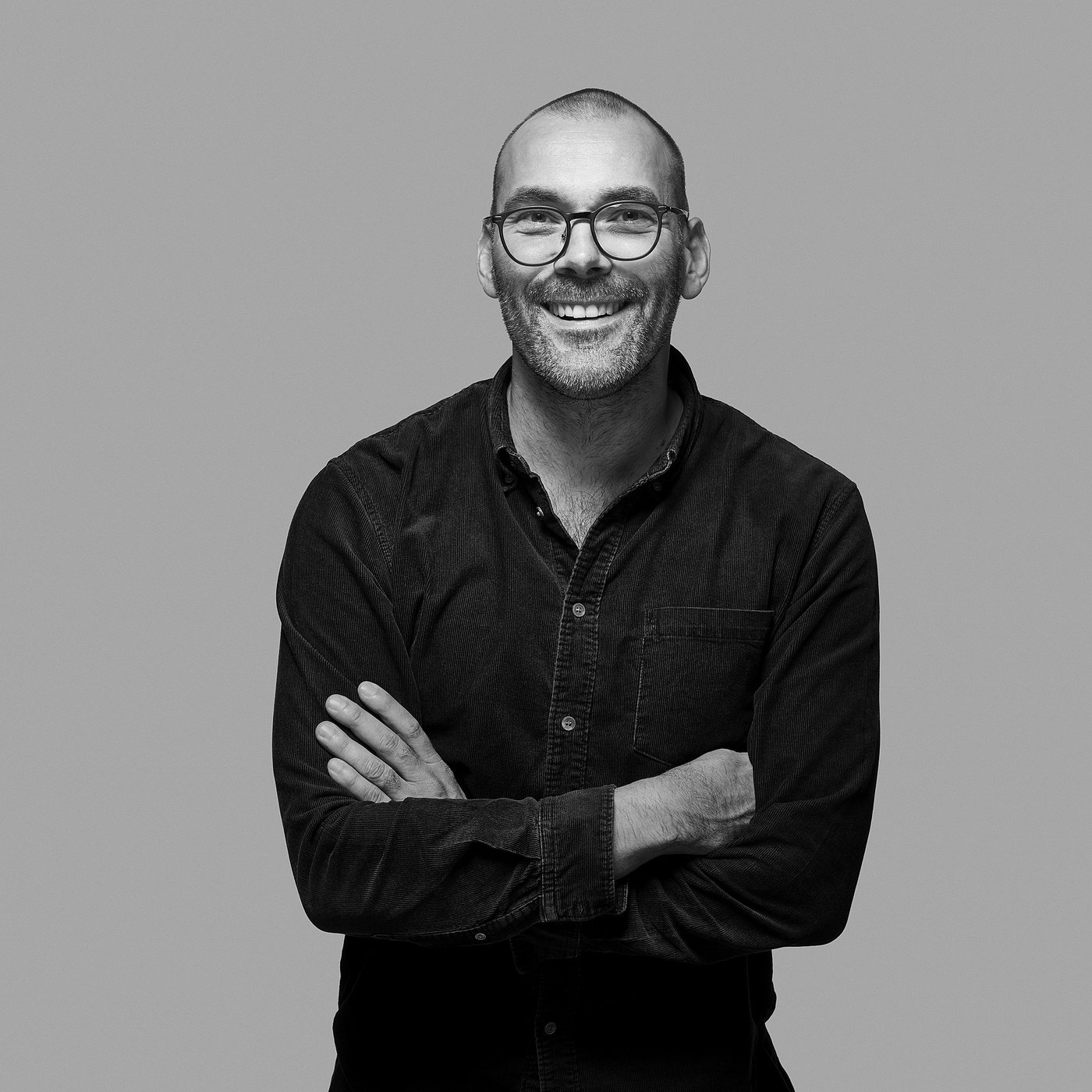 Black-and-white portrait of the smiling designer with glasses, arms crossed, wearing a dark shirt.