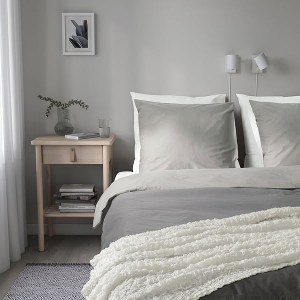 Minimalist bedroom with white bedding, grey pillows, and bedside table holding books, vase, and lamp.