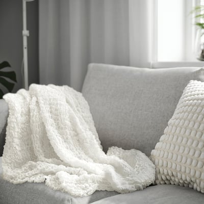 White, soft OFELIA throw blanket on grey couch, light, stretchy, machine washable, easy to carry.