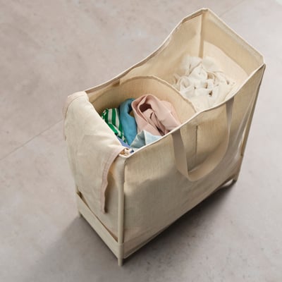 A beige PURRPINGLA laundry basket with a plastic-coated, two-compartment bag. It has sturdy handles and a metal frame for stability.