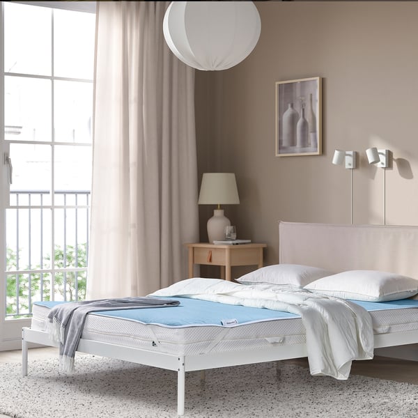 Modern bedroom with a large bed featuring REXBEGONIA blue protector, white linens, wooden bedside table, large window, and minimalist decor.