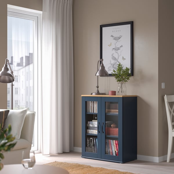 Blue cabinet under art, holding books, next to window and chair.
