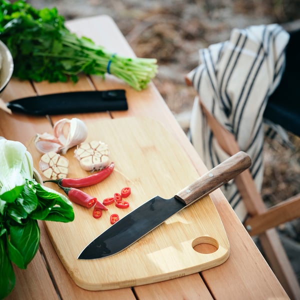 Outdoor kitchen prep with SOLUPPGÅNG knife on wooden board, featuring garlic, chilies, herbs, and striped cloth.