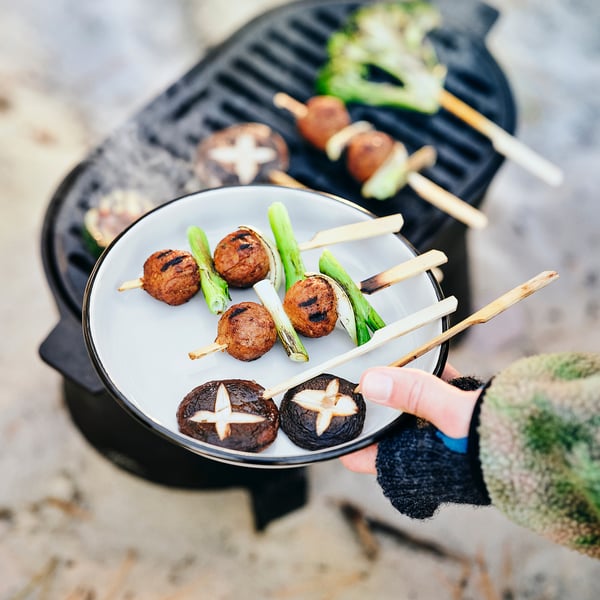 Person holding a plate of grilled skewers with vegetables near a grill. Plate has a rim for better grip, made of lightweight, durable enamel.