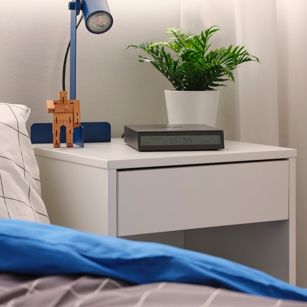 Modern wireless charging alarm clock with temperature, humidity, adjustable backlight. On bedside table with lamp, plant, robot.