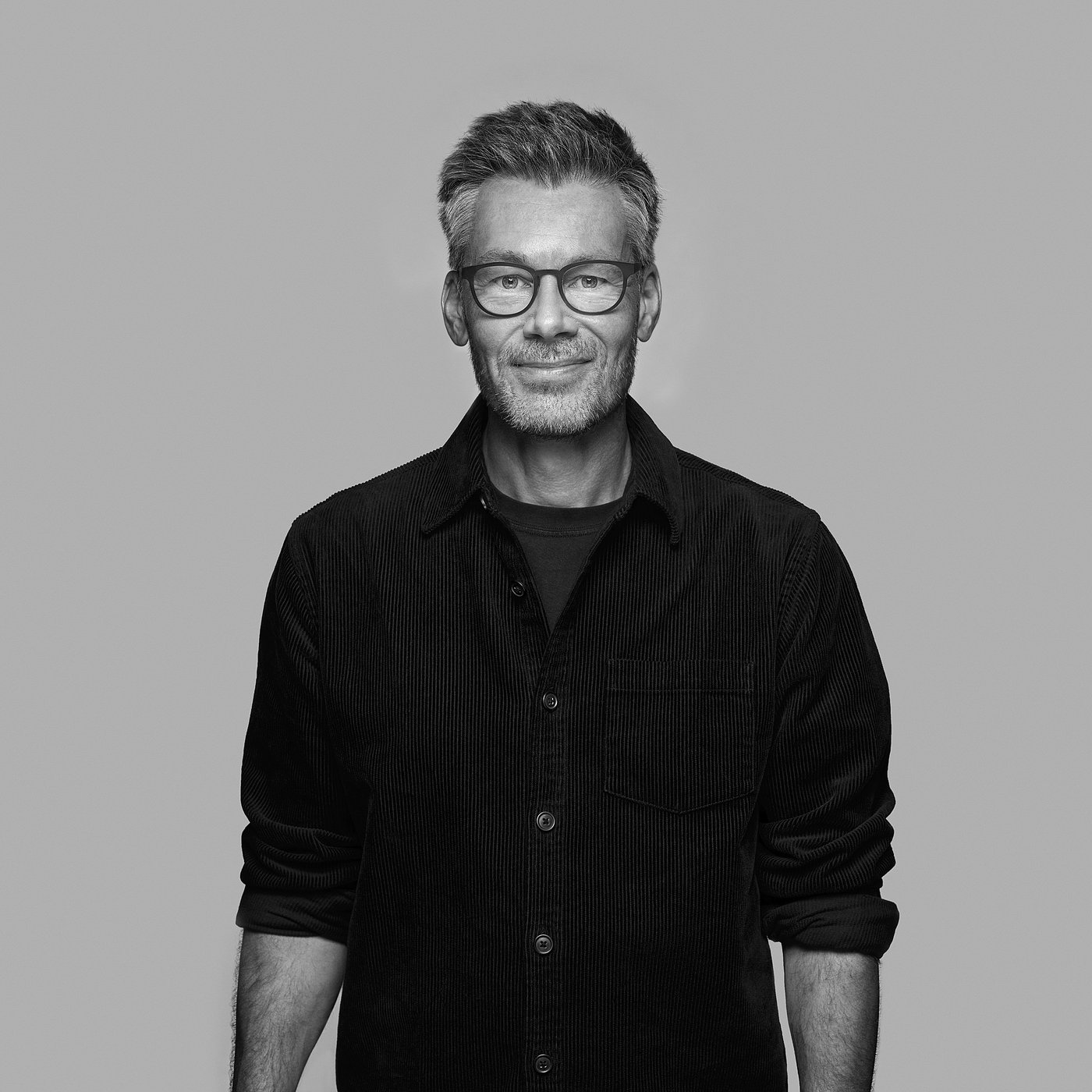 Black-and-white portrait of the designer smiling, wearing glasses and a dark shirt.