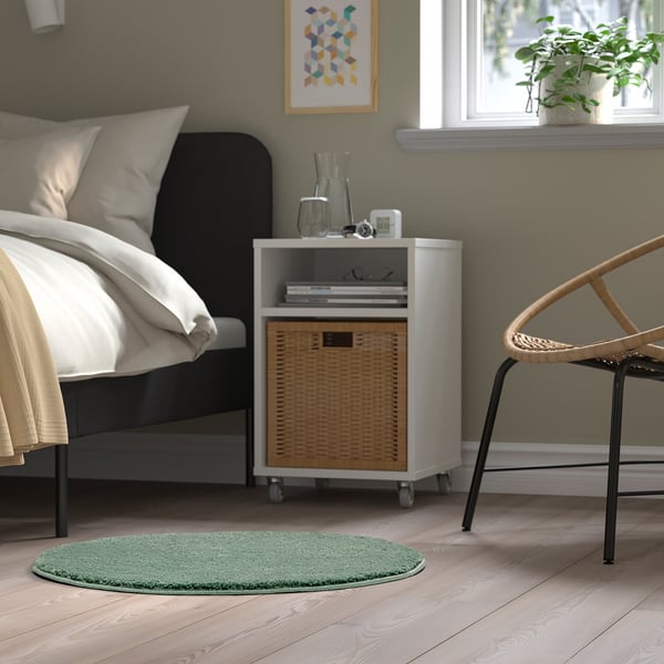 Bedside with green round rug, wicker basket, plant, chair.