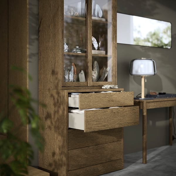 Wooden cabinet with glass doors and open drawers showing items inside, in a room with a lamp.
