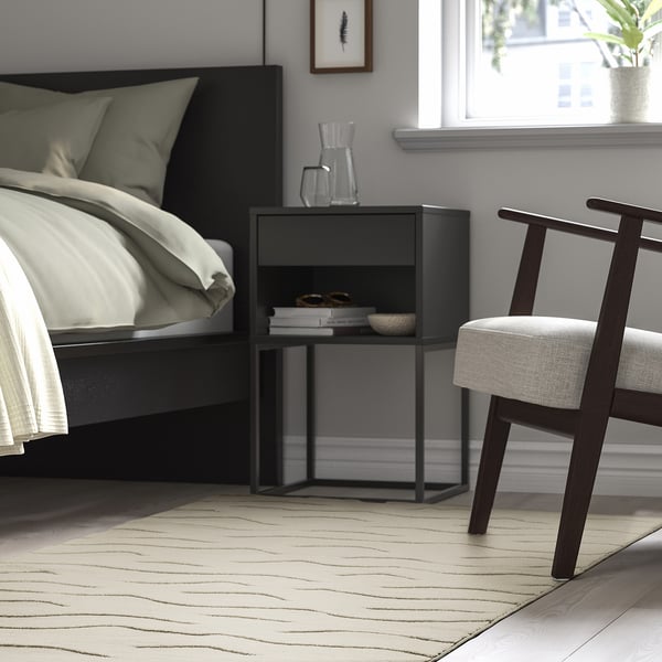 Modern bedroom with black bed, small black side table, white chair, and beige VÄXELFEL rug.